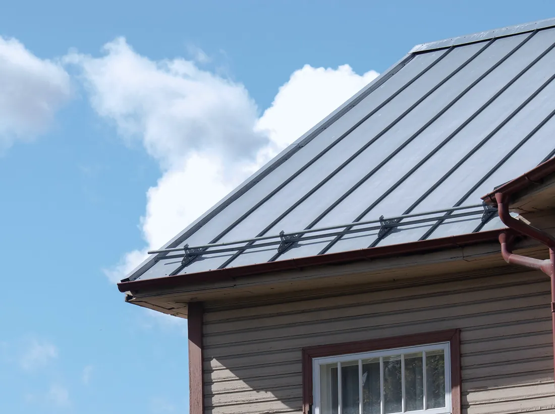 Grey standing-seam metal roof with gutter and downpipe against a blue sky